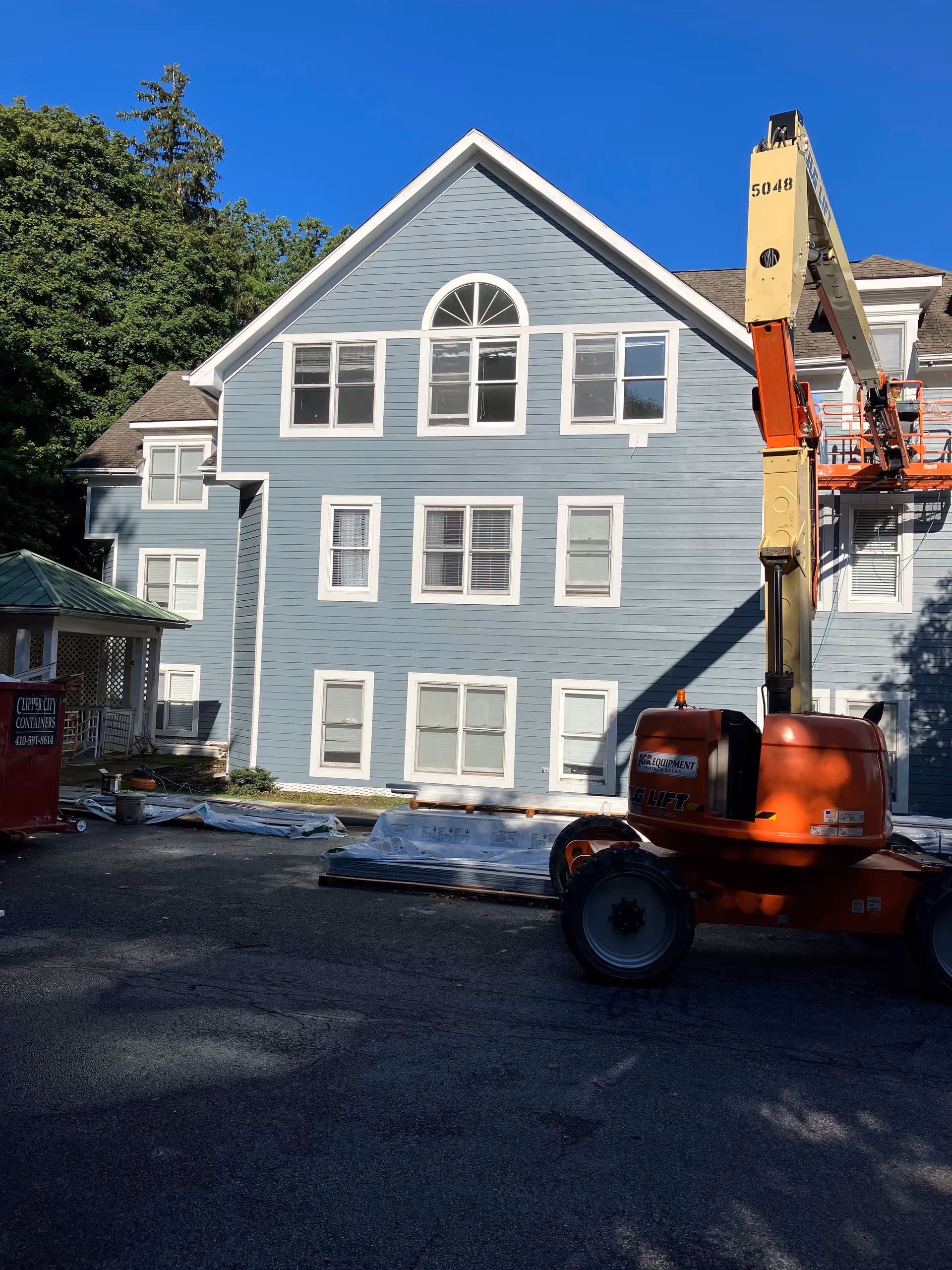 Exterior view of a blue multi-story building with white trim windows under a clear blue sky. There is construction equipment, including an orange lift, and some materials on the ground in front of the building. Trees are visible in the background.