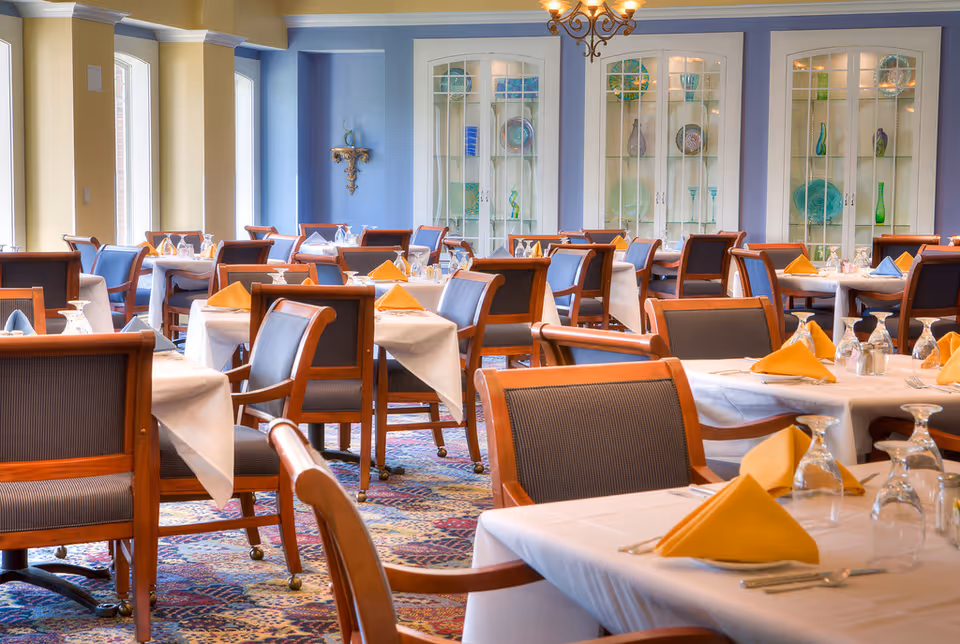 A bright dining room with neatly set tables covered in white linens, yellow napkins, wooden chairs, and glass-front display cabinets in the background.