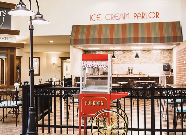 Interior view of a senior living facility area featuring a red popcorn cart with yellow wheels in the foreground. Behind the cart is a black metal fence enclosing a seating area with tables and chairs. Above the seating area is a striped awning with the words 'ICE CREAM PARLOR' written on the wall. To the left, there is a lamp post and a sign for a barber shop.