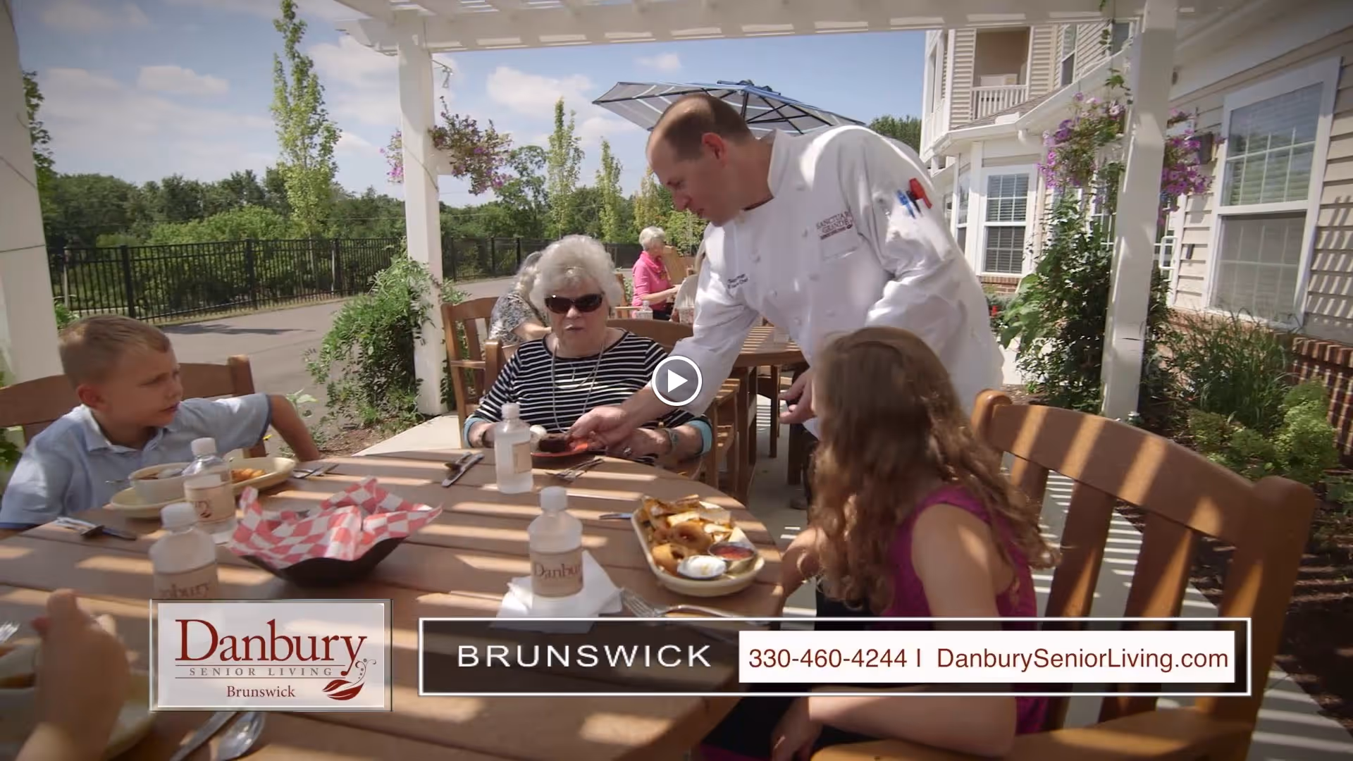 An outdoor patio dining area at Danbury Brunswick with a chef serving food to an elderly woman and two children seated at a wooden table under a pergola. The setting is sunny with greenery and flowers around.
