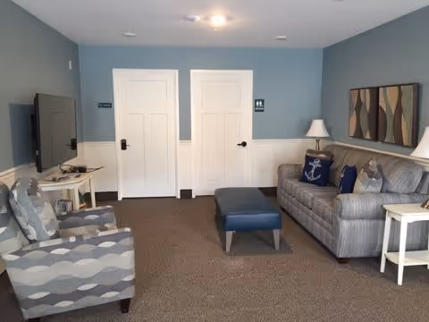 Communal living room with a striped sofa, patterned armchairs, an ottoman, TV on a stand, side tables, and double white doors against blue walls.