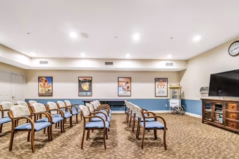 A communal activity room with rows of upholstered chairs arranged theater-style facing a large TV and framed posters on the wall.