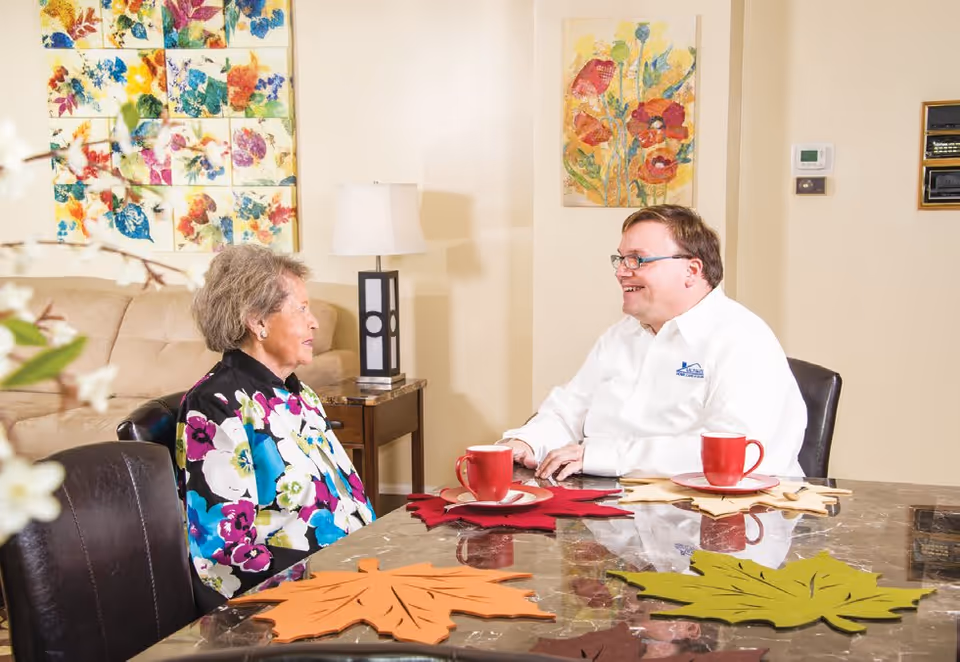 An elderly woman and a man are sitting at a table in a warmly decorated room, engaged in conversation. The table has colorful leaf-shaped placemats and red mugs. Behind them, there is a beige couch, a side table with a lamp, and floral artwork on the walls.