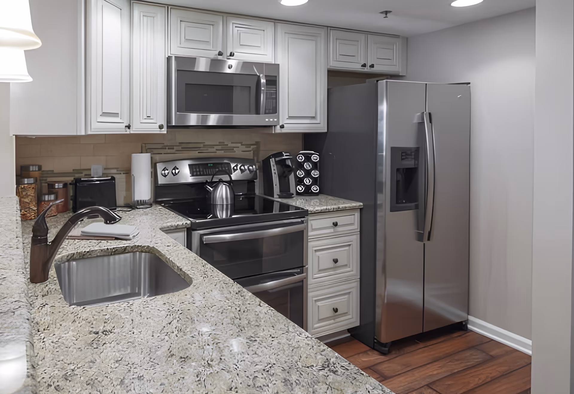 Modern kitchen with granite countertops, stainless steel appliances including a refrigerator, stove, and microwave. White cabinetry and a sink with a bronze faucet are visible. The floor is wooden and the walls are painted light gray.
