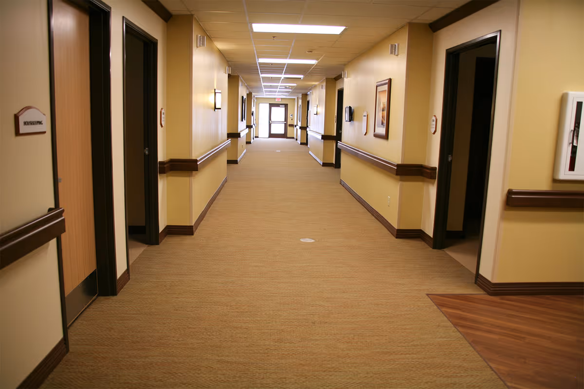 A long, well-lit hallway in a senior living facility with beige walls and carpeted floor. Several doors line both sides of the hallway, with handrails along the walls. There are framed pictures and wall-mounted lights along the corridor, and an exit door is visible at the far end.