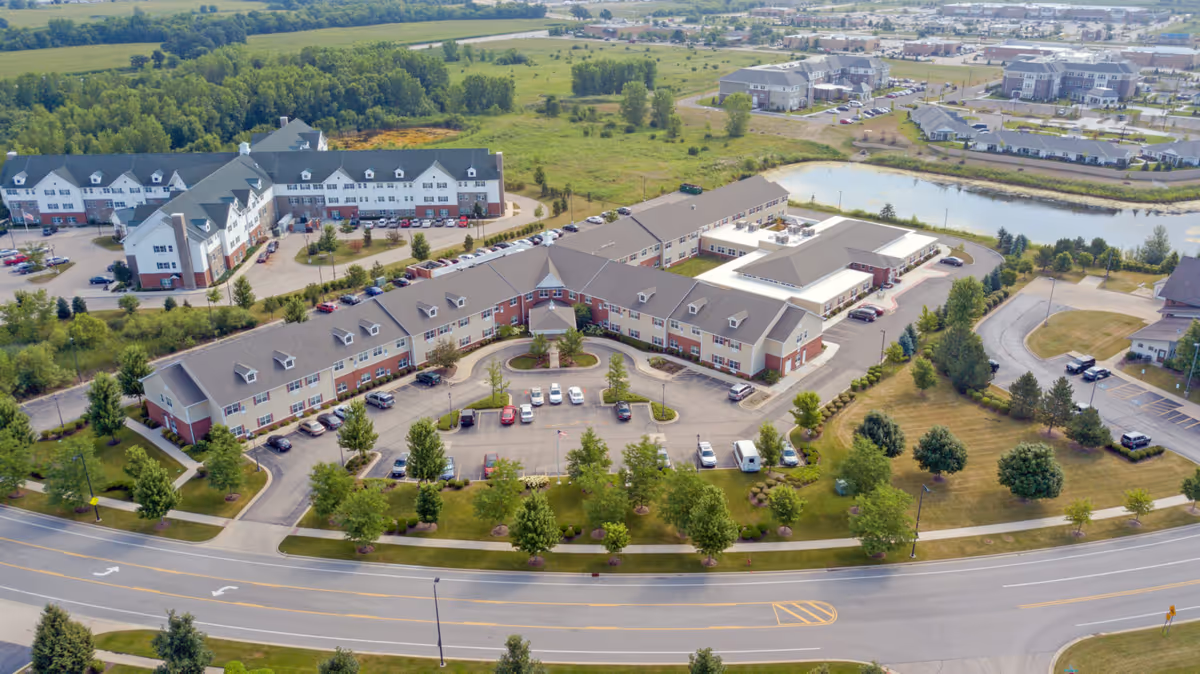 Aerial view of Heritage Woods of Huntley senior living facility showing multiple connected buildings with parking lots, landscaped green areas, trees, and a nearby pond. The facility is surrounded by roads and other buildings in the background.