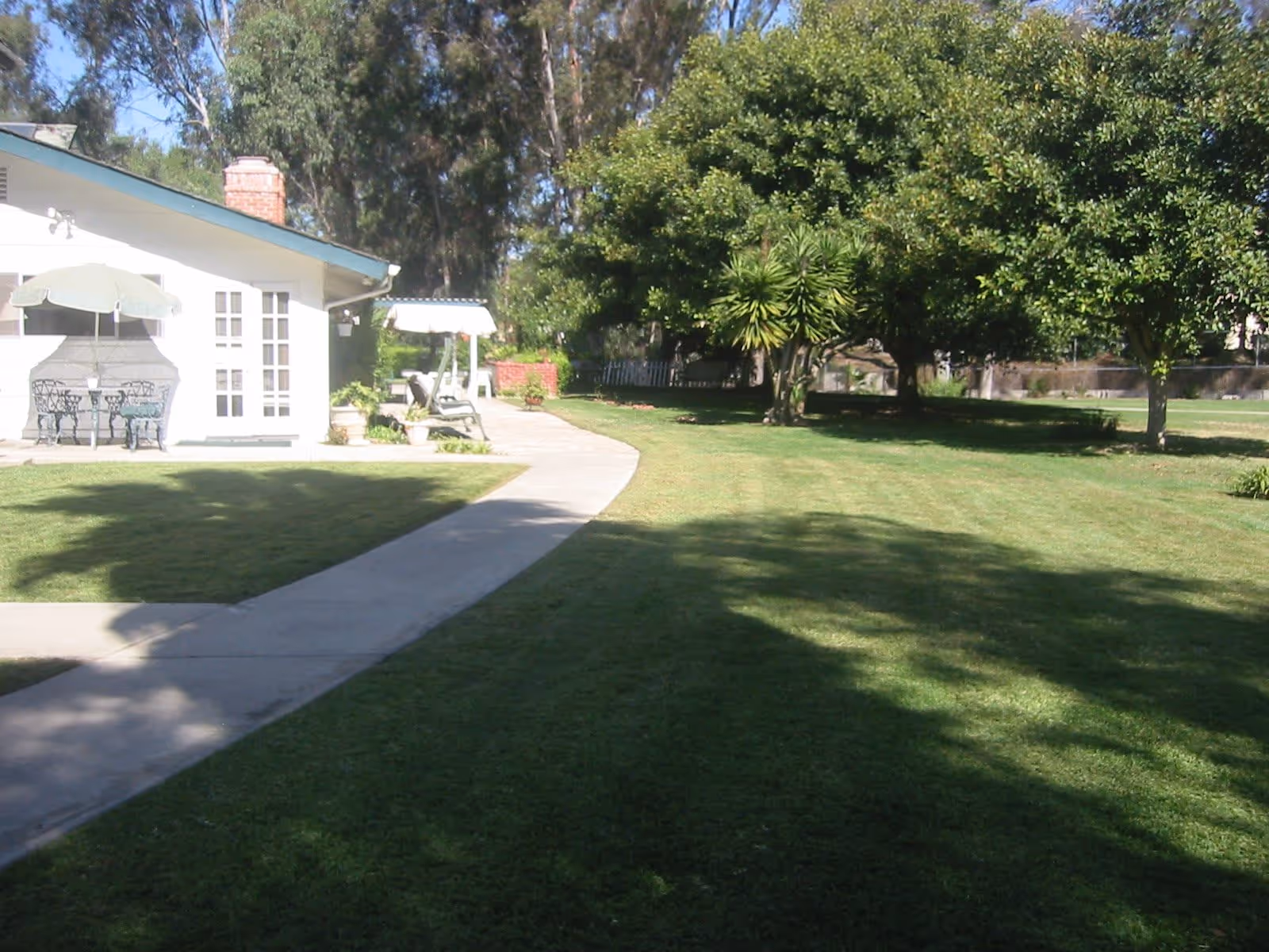 A sunny outdoor garden area at Shadowridge Senior Living featuring a paved walkway curving through a well-maintained lawn with large trees providing shade. To the left, there is a white building with a green roof, a patio table with an umbrella, and a swing bench.