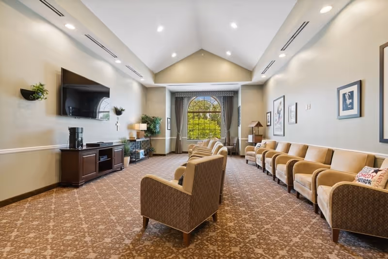 A spacious living room with a high vaulted ceiling, patterned carpet, and beige walls. The room features a row of beige armchairs along one wall and a few additional armchairs facing a large flat-screen TV mounted on the opposite wall. There are framed pictures on the walls, potted plants, a wooden cabinet under the TV, and a large arched window with curtains letting in natural light.
