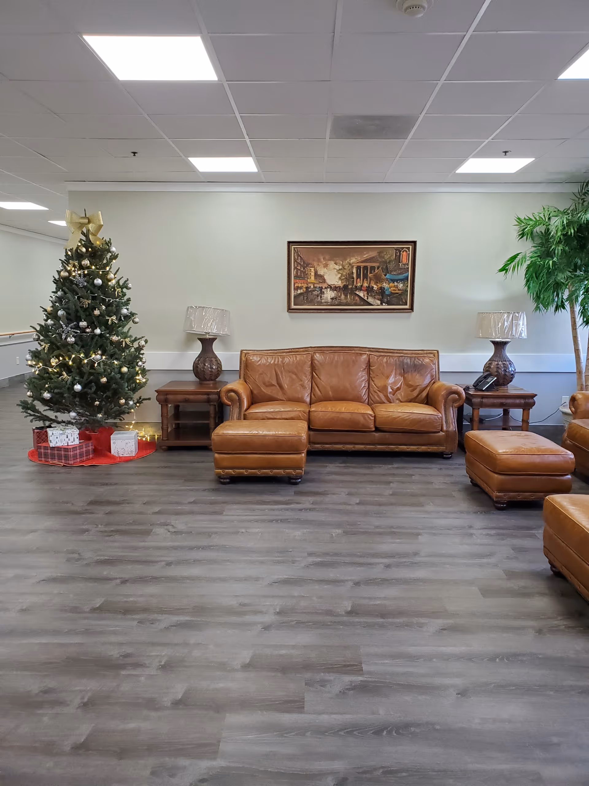 A seating area with a brown leather sofa and ottomans flanked by side tables and lamps, a decorated Christmas tree with gifts, and a framed painting on the wall.
