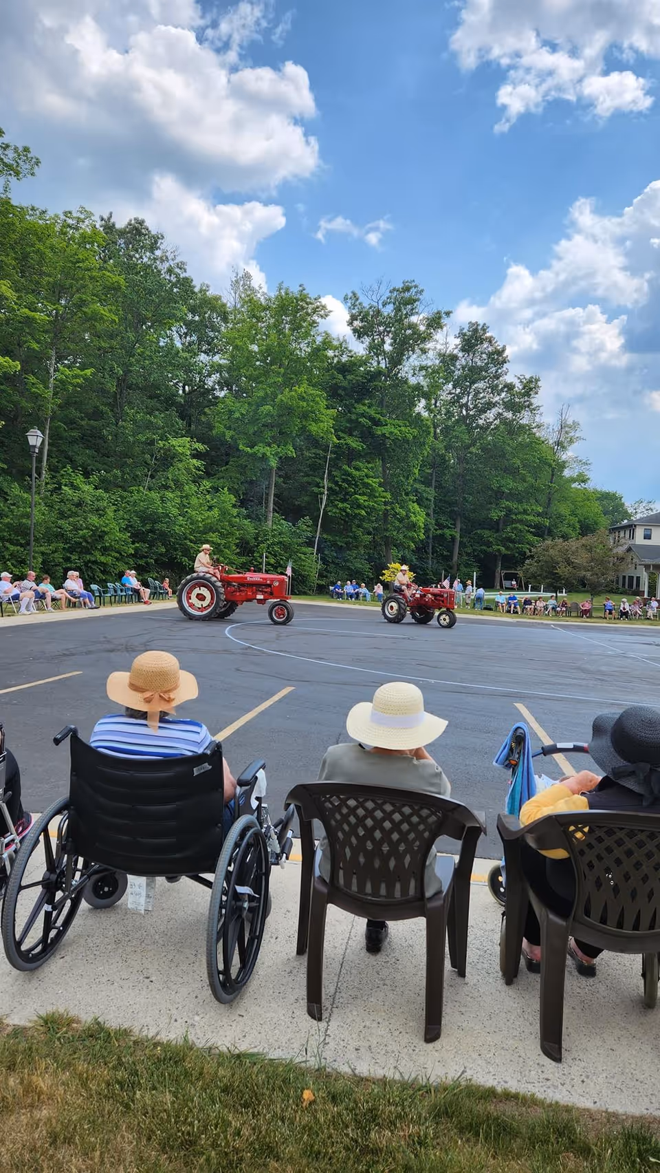 Seniors seated in chairs and wheelchairs watch two vintage red tractors drive in a parking lot with trees and a blue sky.