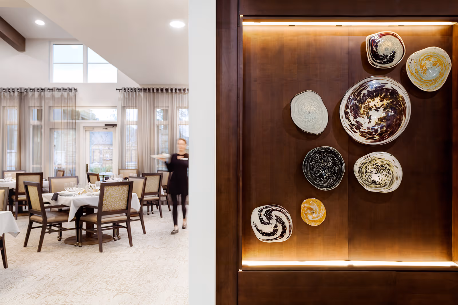 Interior view of a dining room in a senior living facility with tables set with white tablecloths, chairs, large windows with sheer curtains, and a server carrying a plate. On the right side, a wooden wall features decorative ceramic plates mounted as art, illuminated by soft lighting.