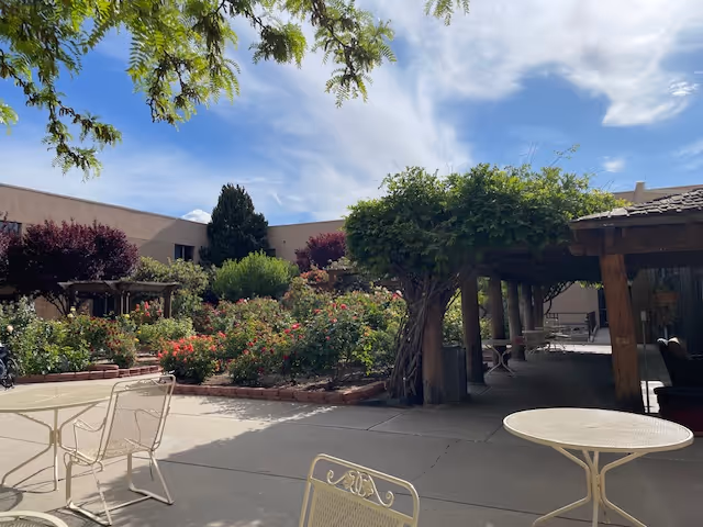 Outdoor patio area with metal chairs and tables, surrounded by lush greenery, flowering bushes, and trees under a partly cloudy sky. A wooden pergola covered with vines provides shaded seating.
