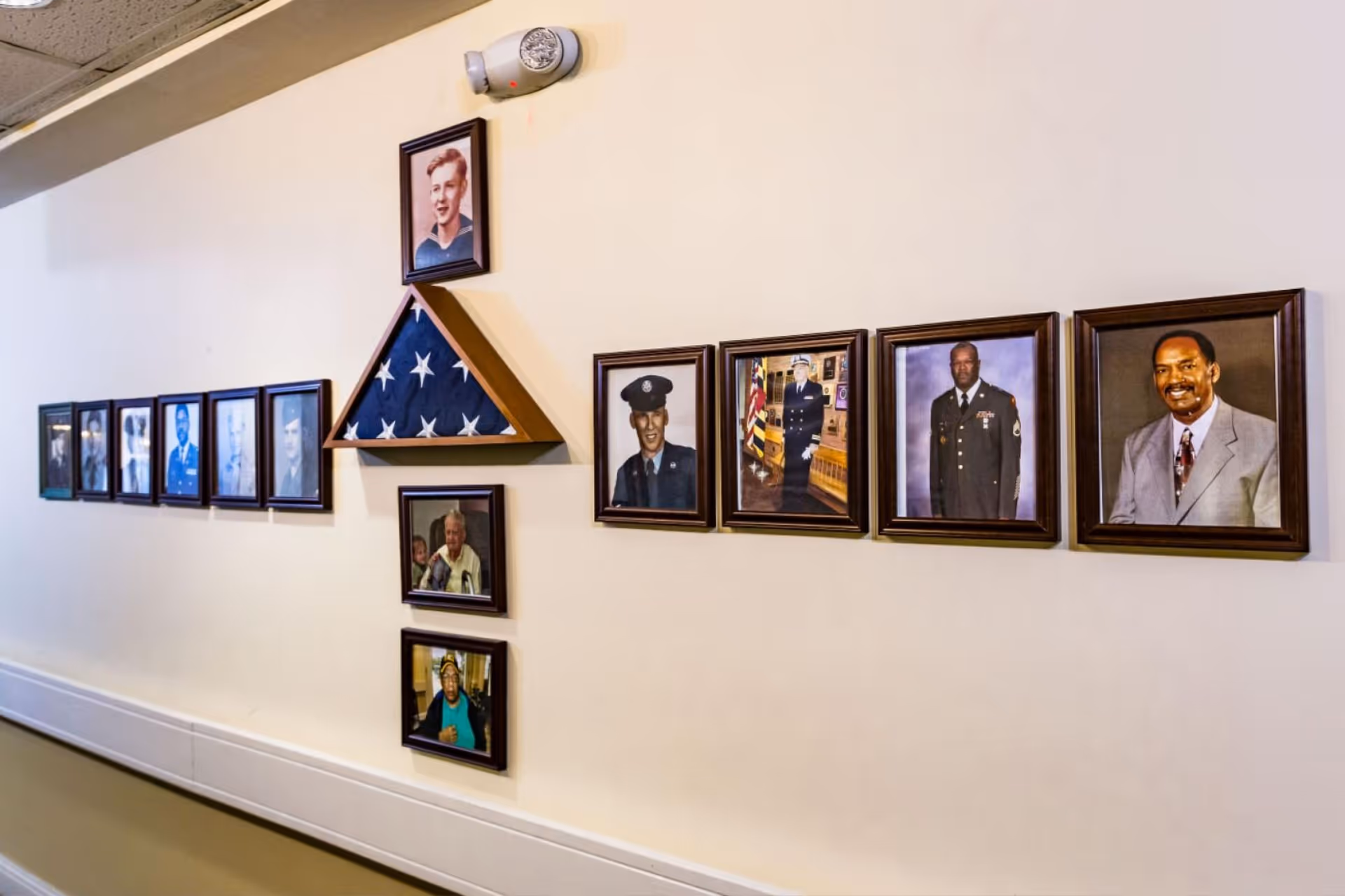 A hallway wall displaying a collection of framed photographs of various individuals, including military personnel in uniform, along with a folded American flag in a triangular wooden case. The wall is painted light beige and has a smoke detector mounted above the flag case.
