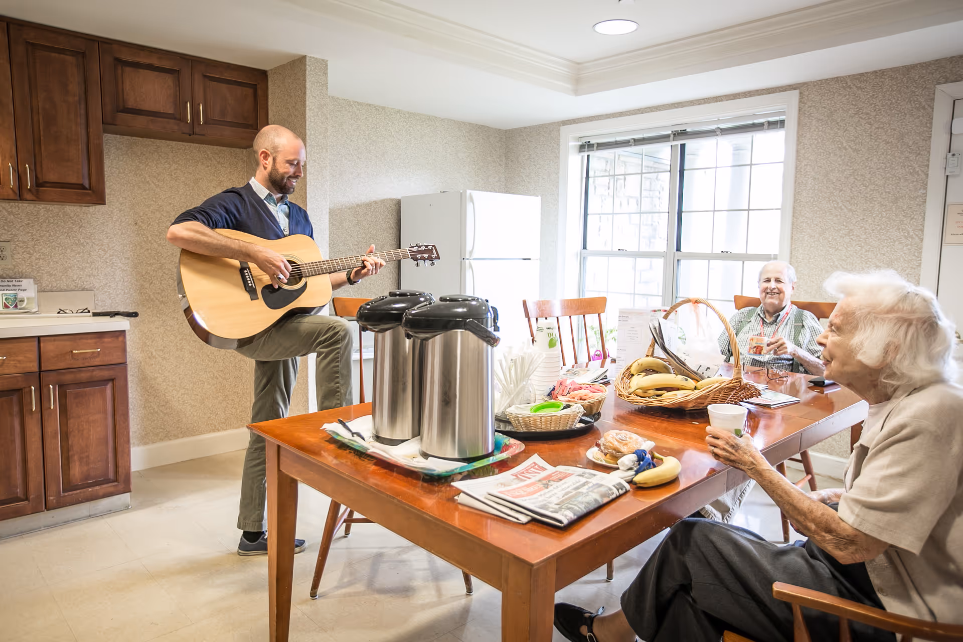 A man playing an acoustic guitar in a kitchen area while two elderly women sit at a wooden table with snacks, bananas, newspapers, and coffee dispensers, enjoying the music and each other's company.
