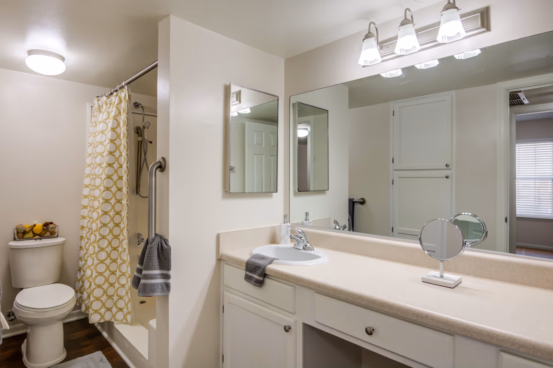 A clean bathroom with a white toilet, a shower with a yellow and white patterned curtain, a large mirror above a beige countertop with a sink, and white cabinets underneath. There are two gray towels hanging, one on the shower wall and one on the countertop. The bathroom has light-colored walls and a wooden floor.