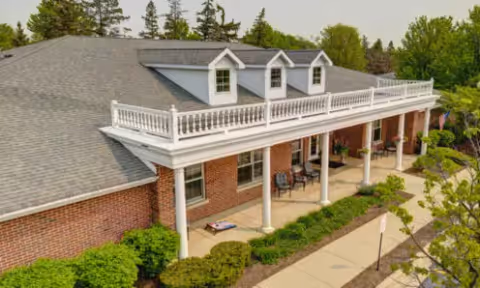 Front exterior of a brick senior living building with a covered porch supported by white columns and an upper balcony, surrounded by shrubs and a walkway.