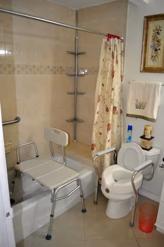 Bathroom with a bathtub equipped with a shower chair and grab bars, a toilet with arm supports, a floral shower curtain, a towel hanging on the wall, and various toiletries on the toilet tank.