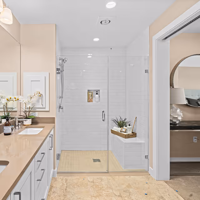 Modern bathroom with a glass-enclosed shower featuring white subway tiles and a built-in bench. The shower has a handheld showerhead and a small recessed shelf with toiletries. To the left, there is a beige countertop with a sink, a potted orchid, and other decorative items. The floor is tiled in a light stone color. An adjacent room with a round mirror and a lamp is partially visible through an open doorway.