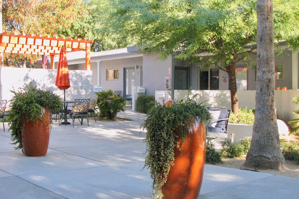 Outdoor courtyard at an assisted living facility with large terracotta planters, patio seating, an orange umbrella, and a low building entrance shaded by trees.