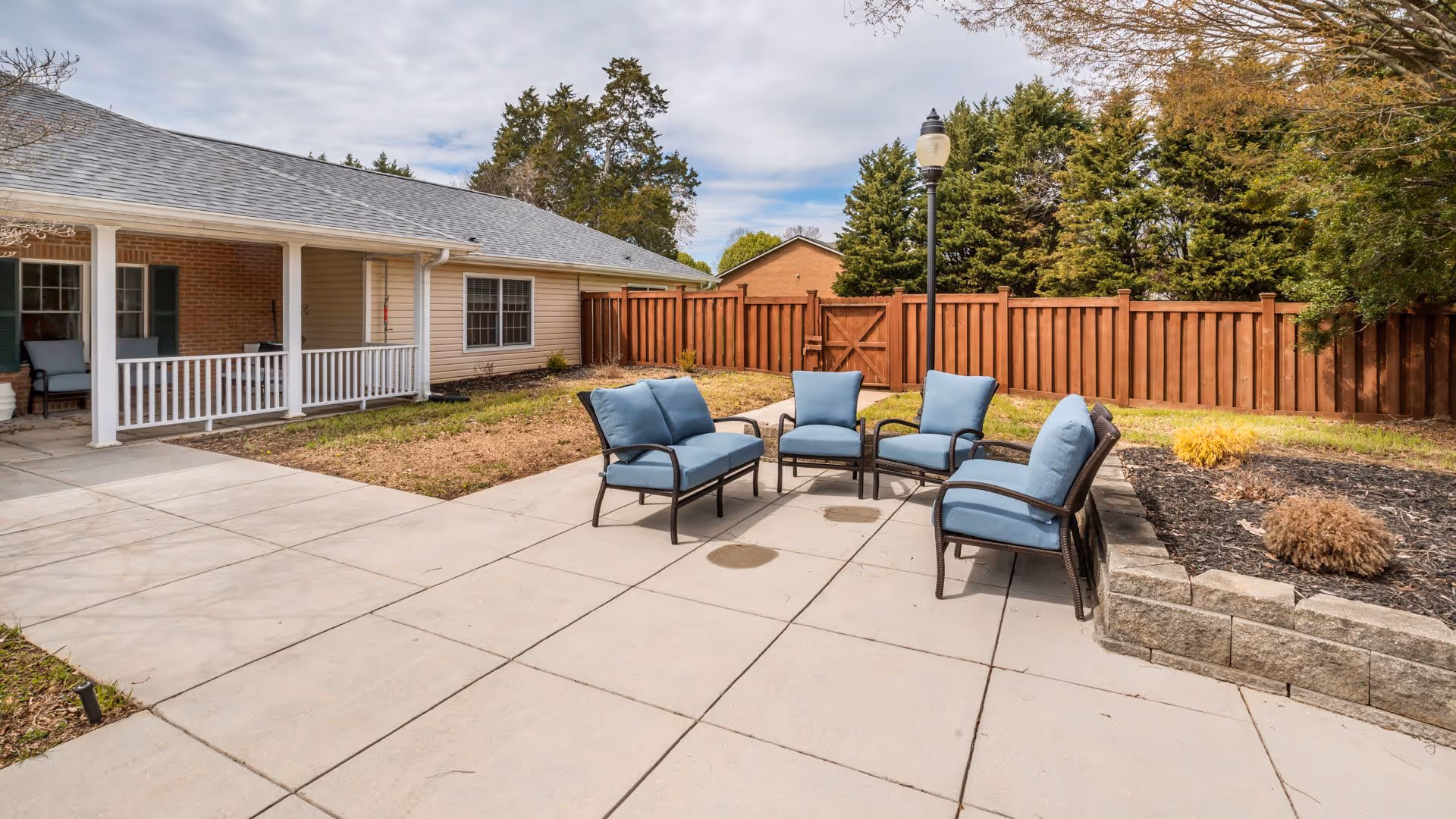 Outdoor patio area at Wellington Manor Knoxville featuring a set of four blue cushioned chairs arranged around a small table on a tiled concrete surface. The patio is adjacent to a single-story building with a covered porch and white railing. A wooden fence and trees surround the area under a partly cloudy sky.