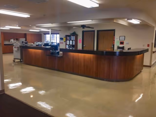 Long wooden reception or nurse station in a well-lit senior living facility interior with beige floors and overhead fluorescent lights.