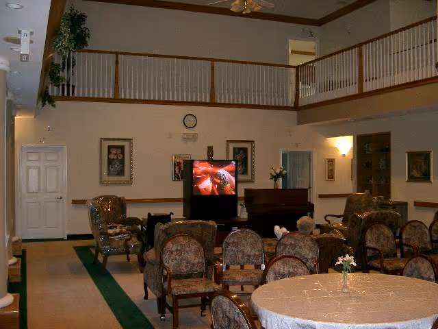 A common area in a senior living facility with multiple upholstered chairs arranged facing a television. The room has a high ceiling with a balcony railing above, framed artwork on the walls, a piano, and a round table with a small flower vase in the foreground. A person is seated watching the TV.