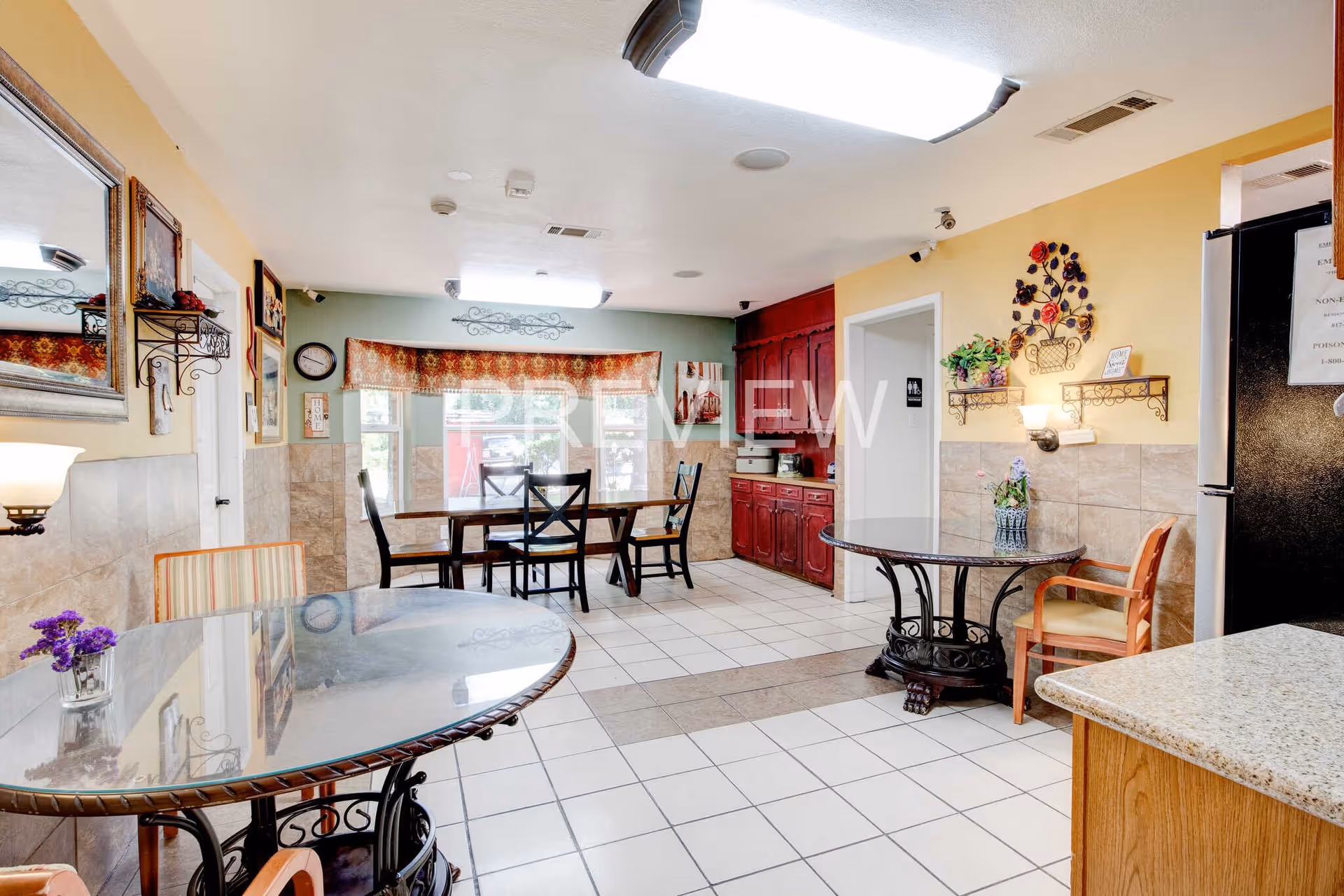 A bright dining area with tiled floors and walls painted in yellow and green. The room features multiple tables with glass tops and chairs arranged around them. There are decorative wall shelves with plants and artwork, a large window with a patterned valance, and a red cabinet with a countertop. A refrigerator is visible on the right side, and a door leads to another room.