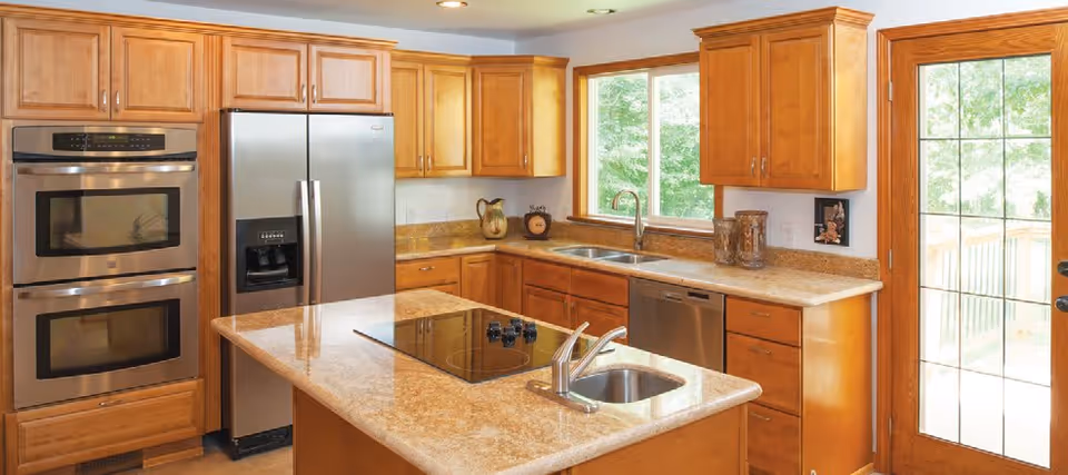 A modern kitchen with wooden cabinets, granite countertops, a double oven, a stainless steel refrigerator, a dishwasher, a kitchen island with a built-in sink and stovetop, and large windows letting in natural light.