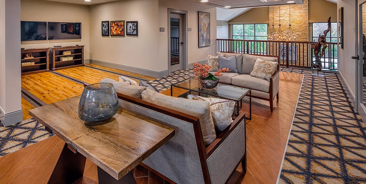 A cozy living room area with two beige sofas arranged around a glass coffee table. A wooden console table with a decorative vase is behind one sofa. The room features patterned carpet and wood flooring, a TV mounted on the wall with a wooden media console below, and framed artwork on the walls. Large windows and a railing overlook a lower level with a stone accent wall and chandelier.