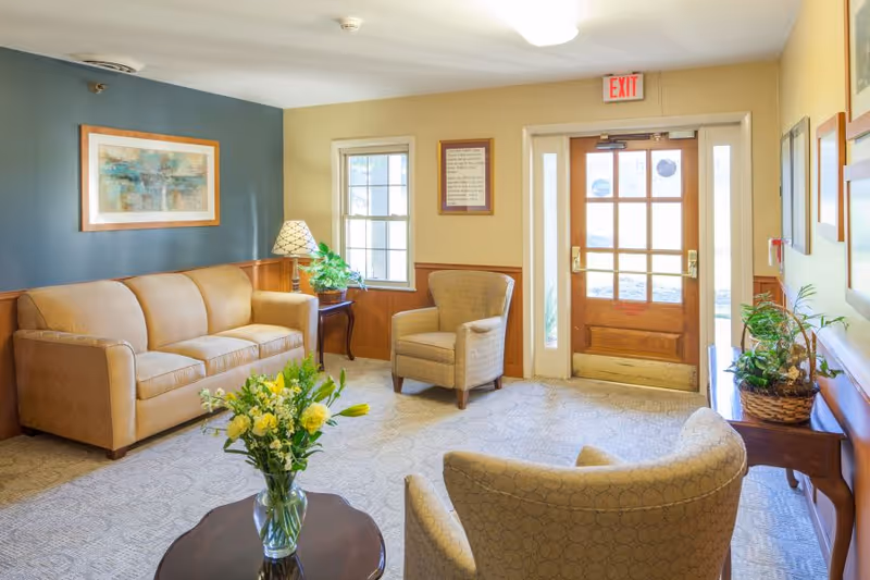 A cozy seating area in a healthcare facility featuring a beige couch, two beige armchairs, a wooden coffee table with a vase of yellow flowers, a side table with a lamp and a plant, framed artwork on the walls, and a wooden door with glass panels leading outside.