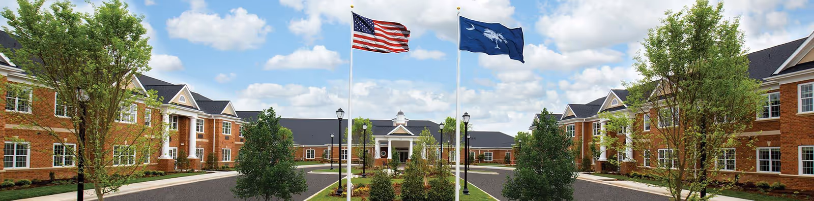 Exterior view of Wellmore of Tega Cay senior living facility showing a large brick building with white columns, two flagpoles with the American flag and South Carolina state flag, trees, and a circular driveway under a partly cloudy sky.