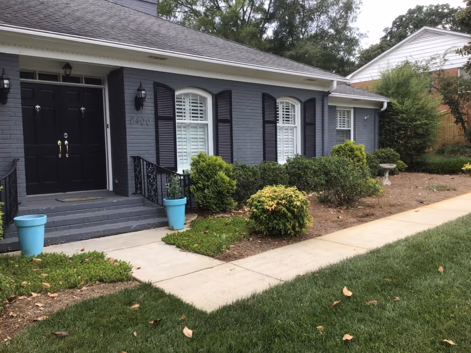 Exterior view of a single-story building with gray brick walls, black double front doors, black shutters on white-framed windows, and a small front porch with steps and black railings. The front yard has green grass, bushes, and two blue flower pots on either side of the steps.