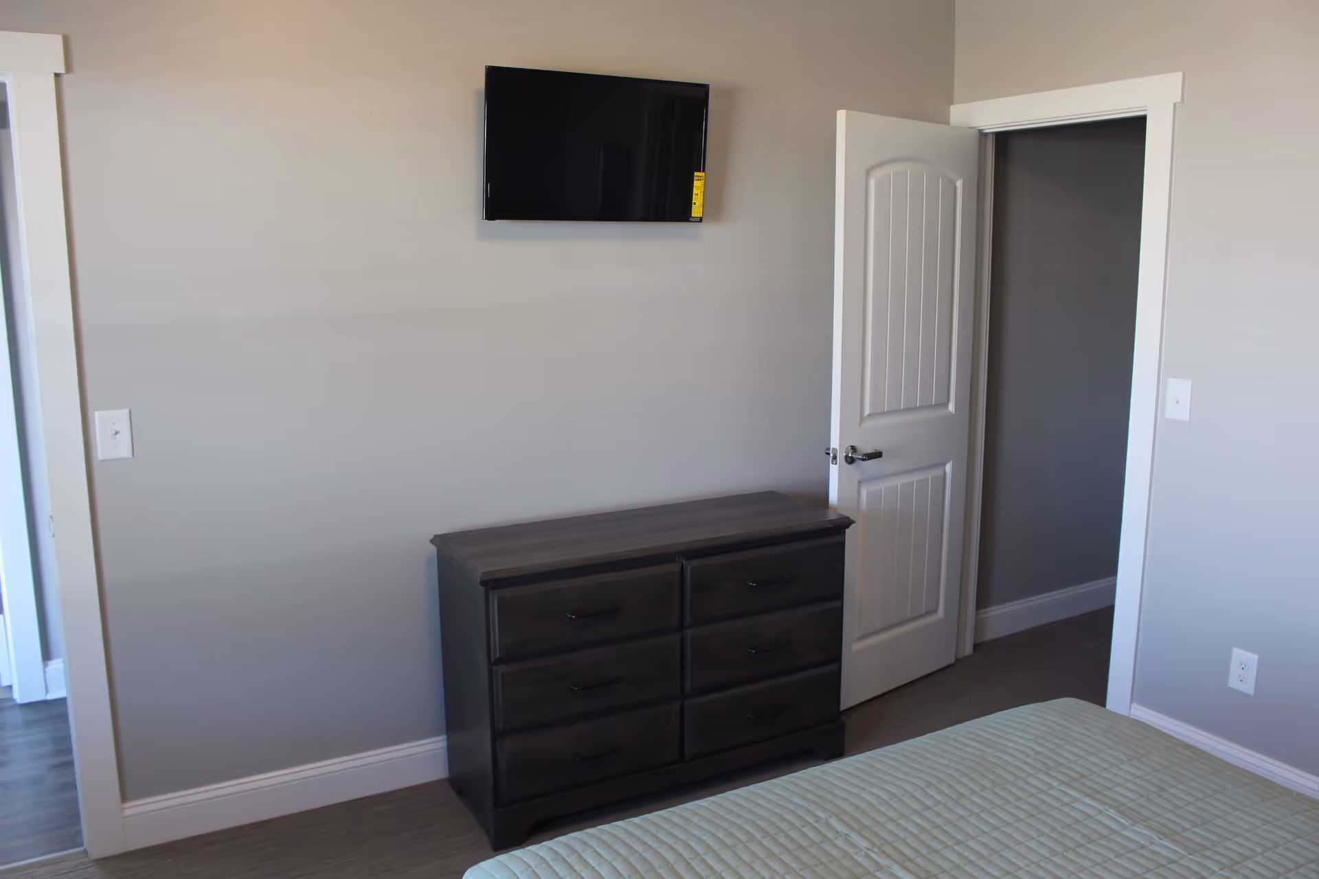 A bedroom with a gray six-drawer dresser against a light gray wall, a flat-screen TV mounted above the dresser, an open white door leading to another room, and a bed with a light green quilted bedspread in the foreground.