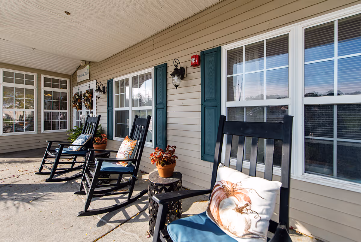 A covered porch area at Whitlock Place with three black rocking chairs, each with cushions and decorative pillows. There are potted plants on small tables between the chairs. The building exterior has beige siding, green shutters, and multiple windows with white frames. A sign above the door reads 'Whitlock Place'.