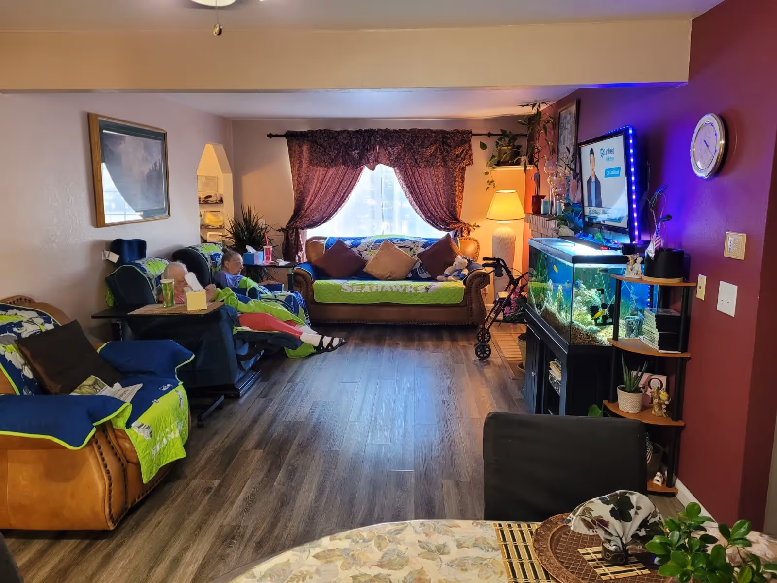 A cozy living room in an adult family home with two elderly people seated in recliners covered with Seahawks-themed blankets. The room features a brown leather sofa with pillows, a large window with purple curtains, a floor lamp, a fish tank with blue LED lights, a wall-mounted TV, and a round dining table with a floral tablecloth in the foreground. The floor is wood laminate, and there are various plants and decorative items around the room.