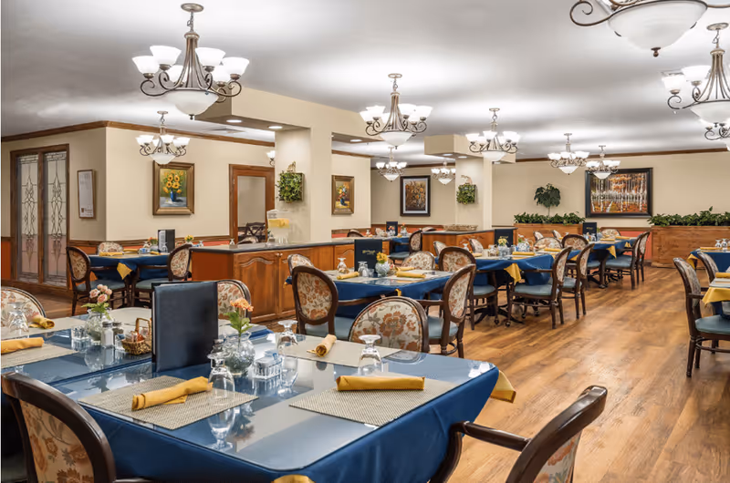 A spacious dining room with multiple tables covered in blue tablecloths and beige placemats, each set with glasses, napkins, and small flower arrangements. The room features wooden flooring, floral upholstered chairs, decorative chandeliers, framed artwork on the walls, and some greenery along the perimeter.