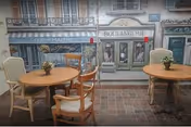 Interior view of a dining area with two round wooden tables, each surrounded by chairs. Small flower arrangements are placed on each table. The background features a mural painted to resemble a quaint street scene with storefronts labeled 'Coffee Masters' and 'Boulangerie'.
