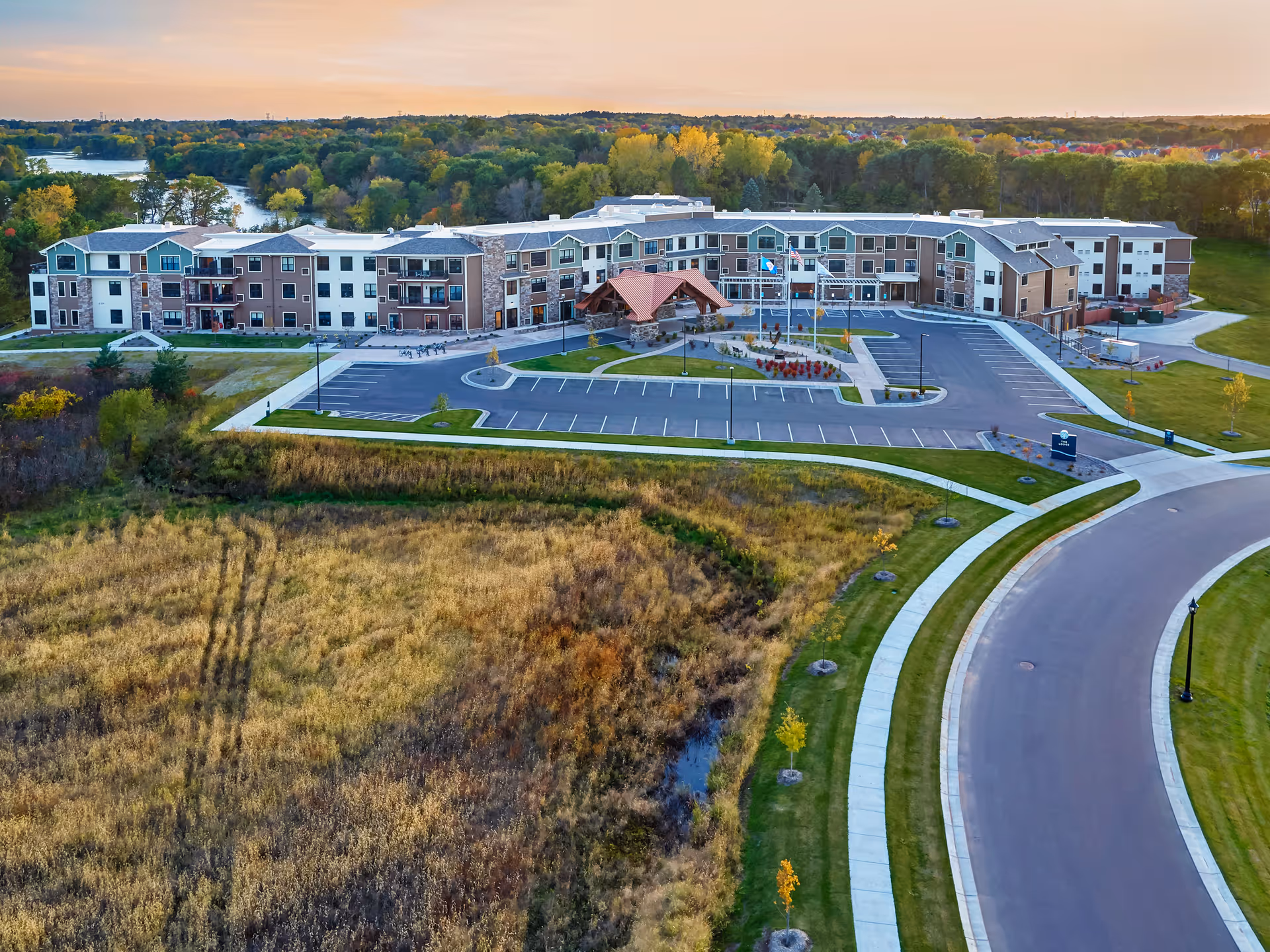Aerial view of The Lakes at Stillwater senior living facility surrounded by greenery and trees with a river in the background, showing a large multi-story building with a parking lot and curved road in front during sunset.
