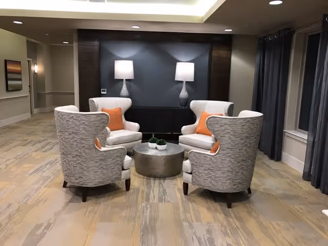 A cozy seating area with four upholstered armchairs arranged around a round metallic coffee table. Each chair has an orange throw pillow. Behind the chairs is a dark accent wall with two table lamps providing soft lighting. The room has a neutral color palette with beige flooring and dark curtains on the right side.