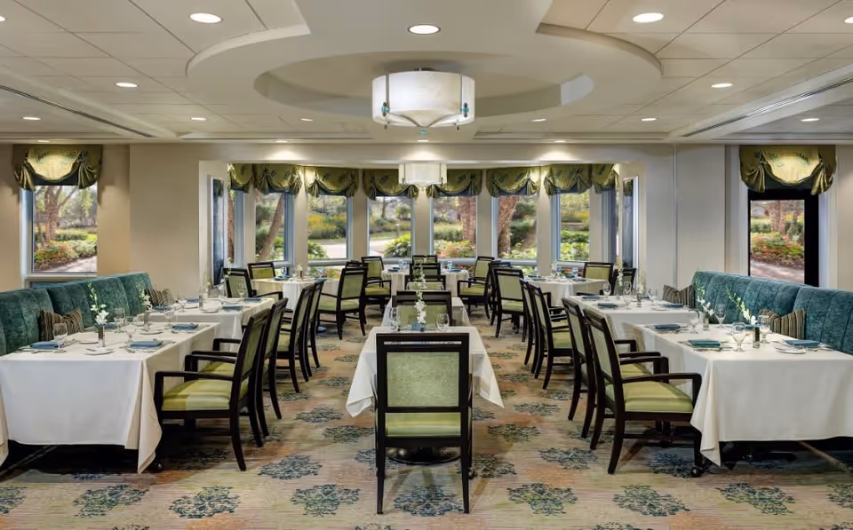 A well-lit dining room with multiple tables covered in white tablecloths, set with plates, glasses, and napkins. Green cushioned chairs surround the tables, and large windows with green valances offer a view of a garden outside. The ceiling features recessed lighting and a central decorative light fixture.