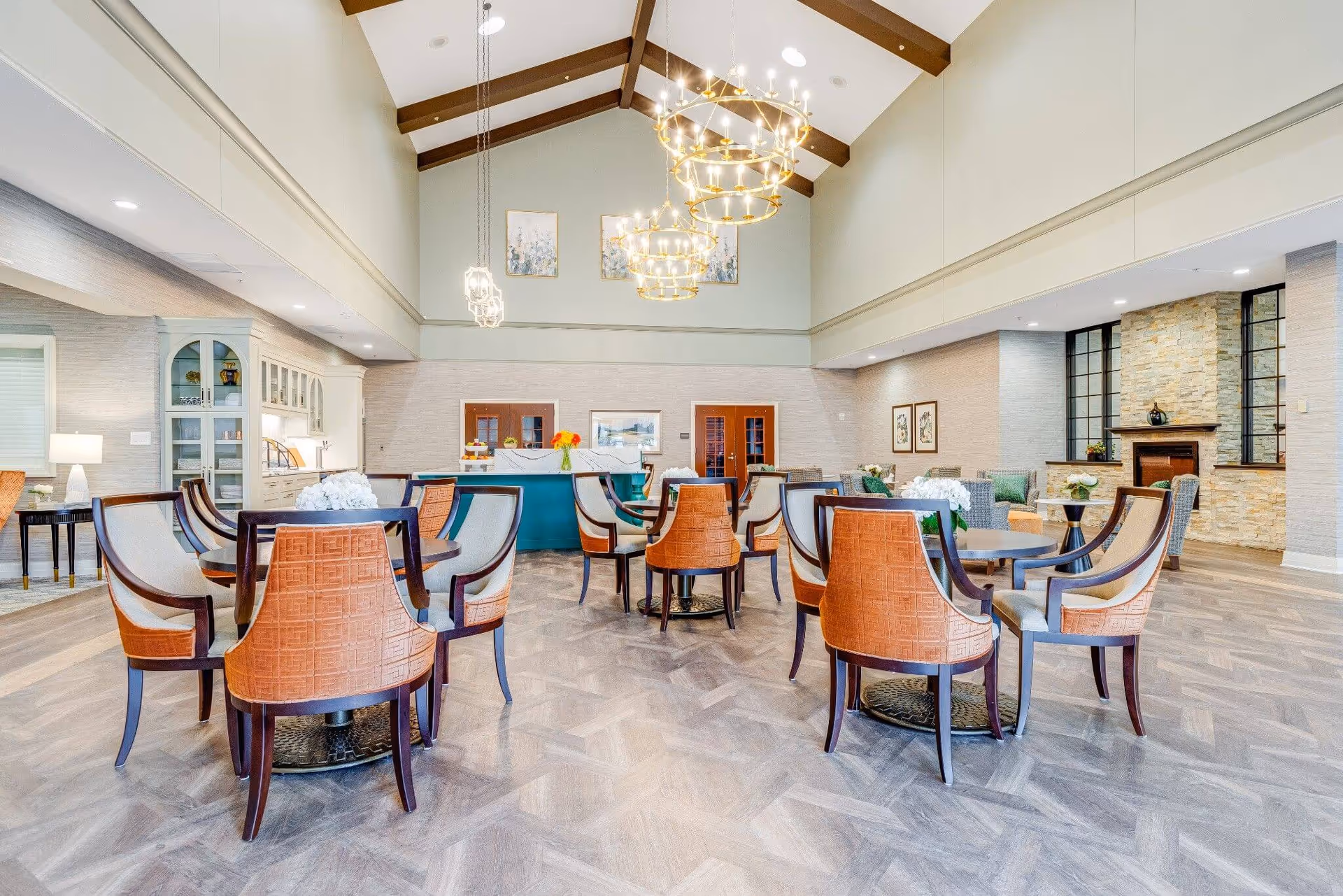 Spacious communal dining and lounge area with round tables, upholstered chairs, high beamed ceiling and chandeliers.
