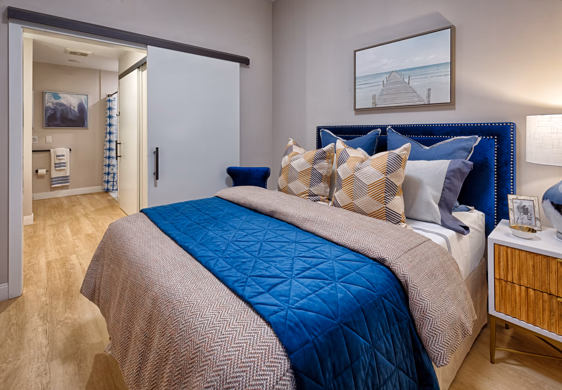 A cozy bedroom with a bed featuring a blue quilt and multiple patterned pillows. A nightstand with a lamp and framed photo is beside the bed. A framed picture of a pier over water hangs above the headboard. In the background, a sliding door leads to a bathroom with a shower curtain and towel rack.