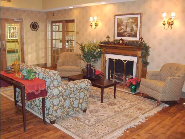 A cozy living room area with a floral patterned sofa, two beige armchairs, a wooden coffee table, and a fireplace decorated with greenery and red poinsettias. The room has hardwood floors, a large patterned rug, wall sconces with lights, a framed picture above the fireplace, and a wooden console table with decorative items and a red runner.