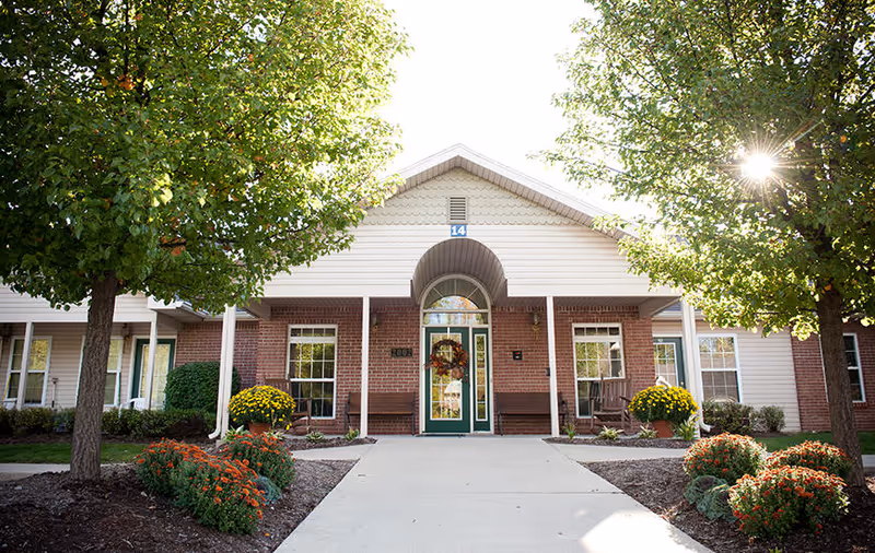 Front exterior view of a single-story building with a peaked roof, brick and siding facade, green door decorated with a wreath, flanked by windows and benches. The entrance is framed by two trees and landscaped flower beds with orange and yellow flowers.