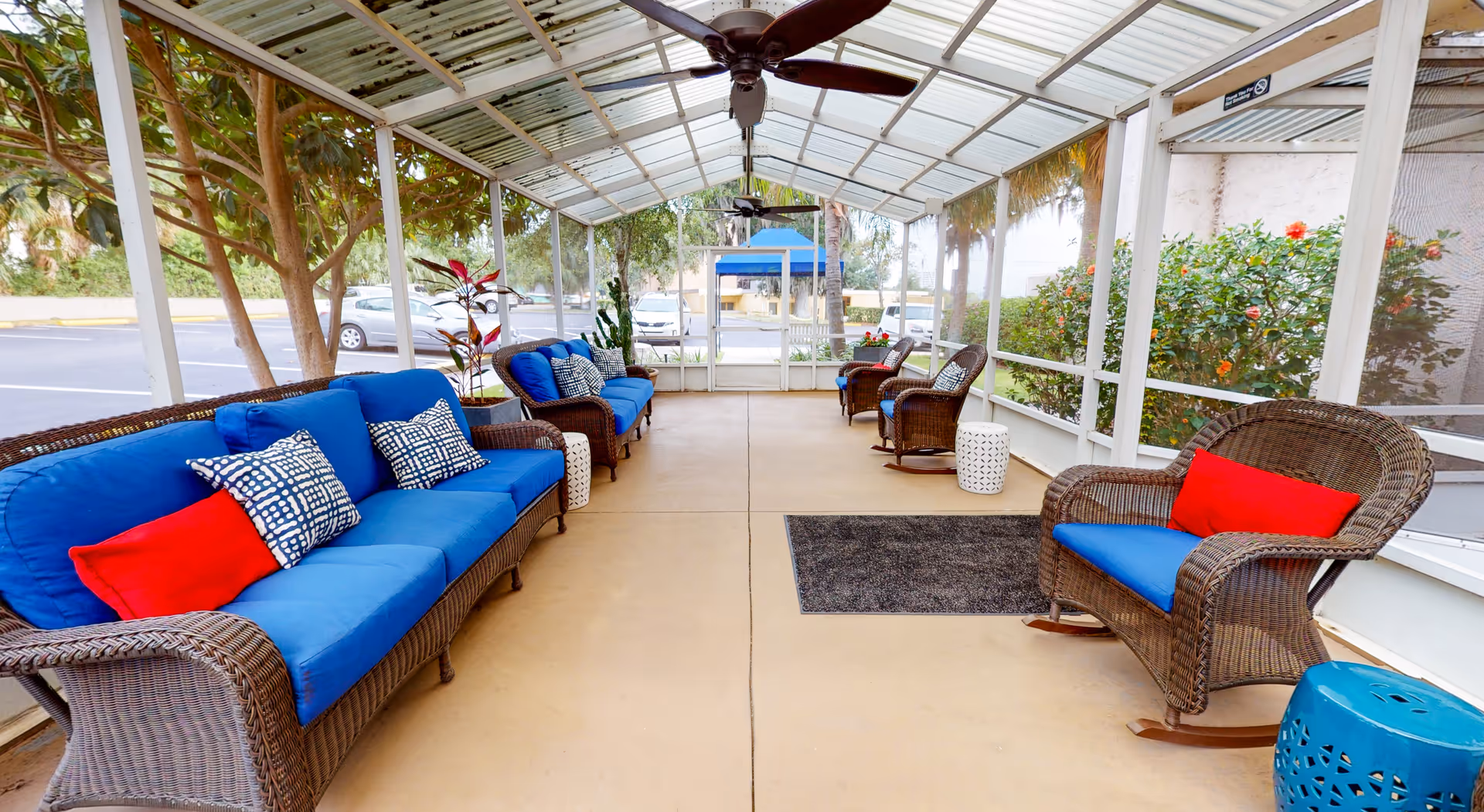 A covered outdoor seating area with wicker furniture featuring blue cushions and red and patterned pillows. The space has ceiling fans, a concrete floor, and is surrounded by screened walls with plants and trees visible outside.