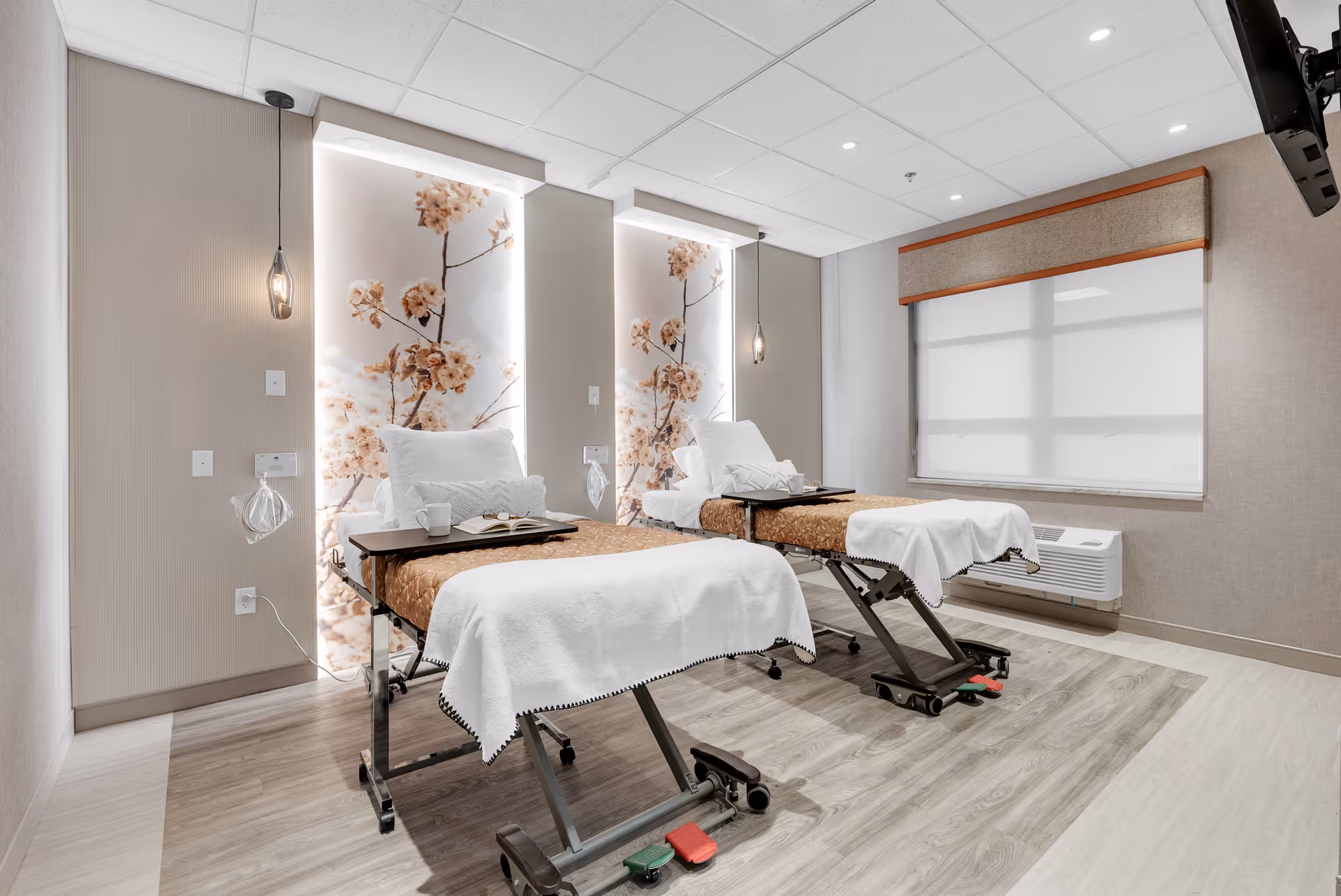 Well-lit rehabilitation-style room with two adjustable hospital beds, floral backlit wall panels, bedside trays, and a large window.