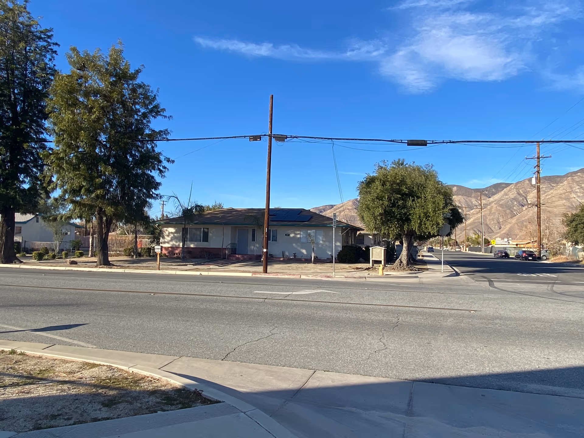 Street view of a single-story building with a few trees around it and mountains in the background under a clear blue sky.