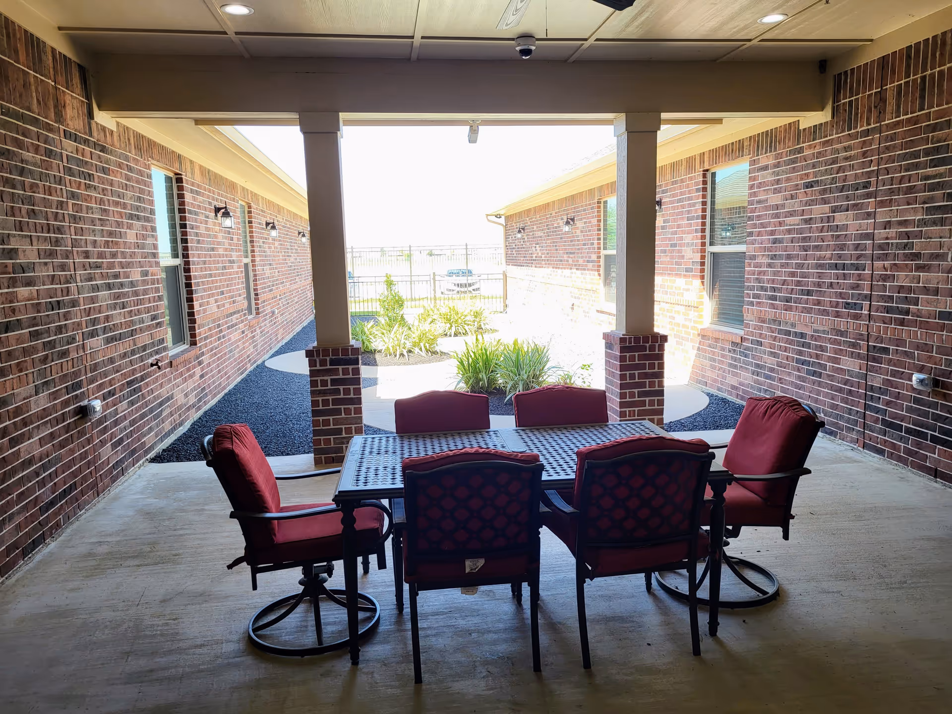 Covered outdoor patio area with a metal table and six red cushioned chairs. The patio is surrounded by brick walls and opens to a small garden area with plants and gravel. The space is shaded by a ceiling with recessed lighting.