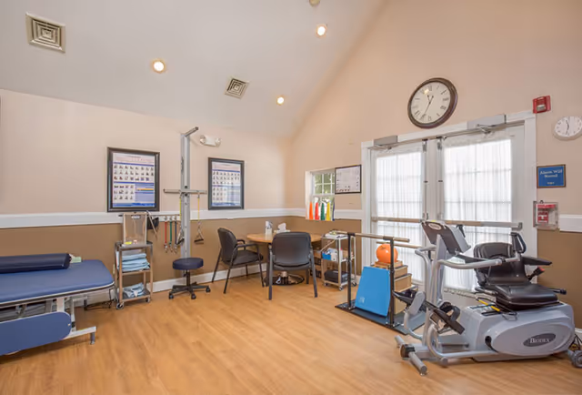 A physical therapy or rehabilitation room with exercise equipment including a stationary bike and parallel bars. There is a treatment table on the left, a small round table with chairs in the center, and medical posters on the walls. The room has wood flooring and a high ceiling with recessed lighting.