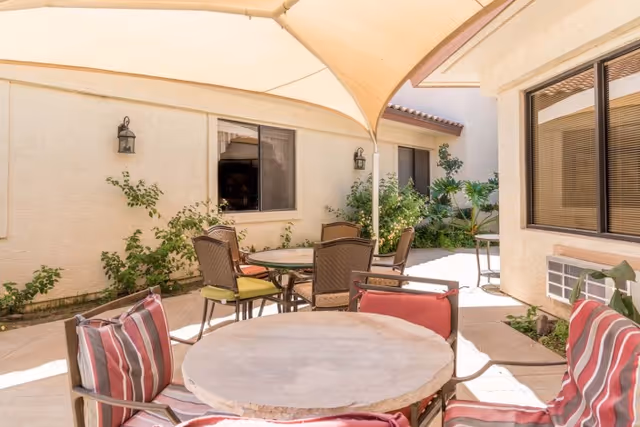 Outdoor patio area with round tables and cushioned chairs under a beige canopy. The patio is surrounded by beige walls with windows and some green plants along the edges.